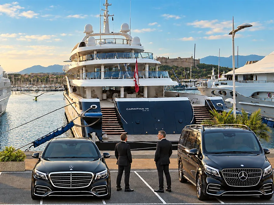 Two chauffeurs in suits wait on the quay beside a black Mercedes-Benz V-Class and a black Mercedes-Benz sedan, in front of moored superyachts, ready to drive owners, guests and crew