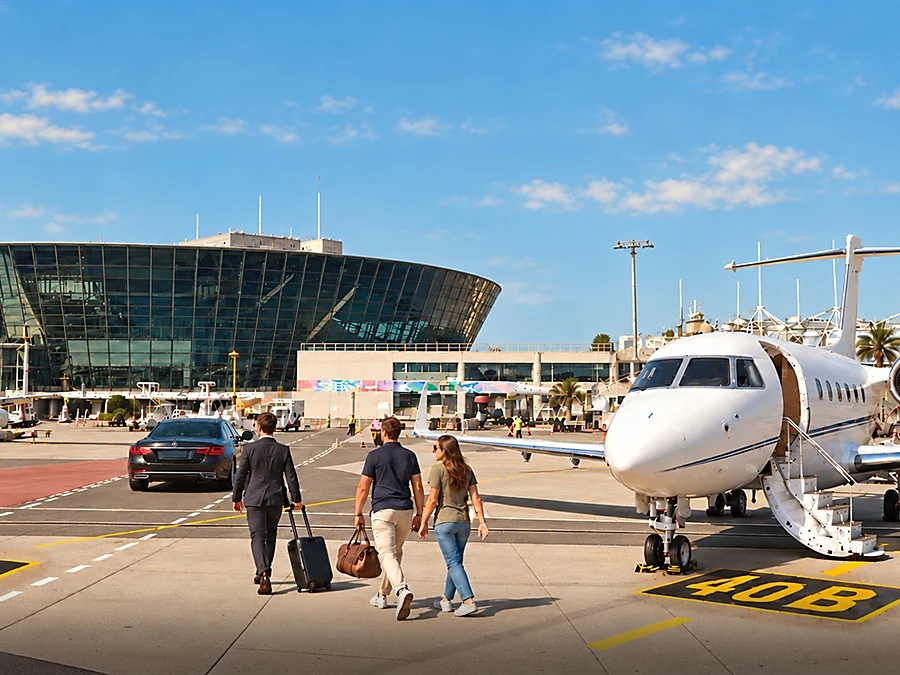 A chauffeur in a suit takes the luggage of a couple arriving by private jet and leads them across the airport apron to a black Mercedes-Benz S-Class