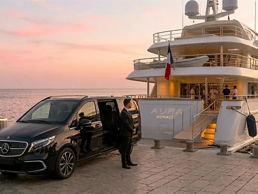 A chauffeur in a suit stands beside a black Mercedes-Benz V-Class on the quay next to a superyacht, ready to drive owners, guests and crew